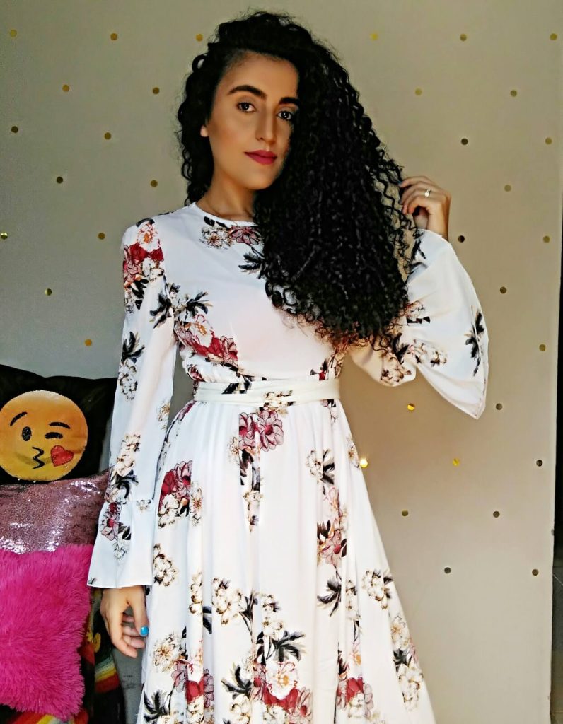 Mulher em vestido floral posa para a câmera, exibindo um sorriso radiante e um fundo natural encantador.